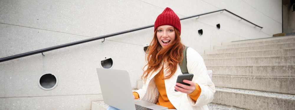 Women in beanie smiling while holding mobile phone and working on laptop at stairs, focused on image protection.