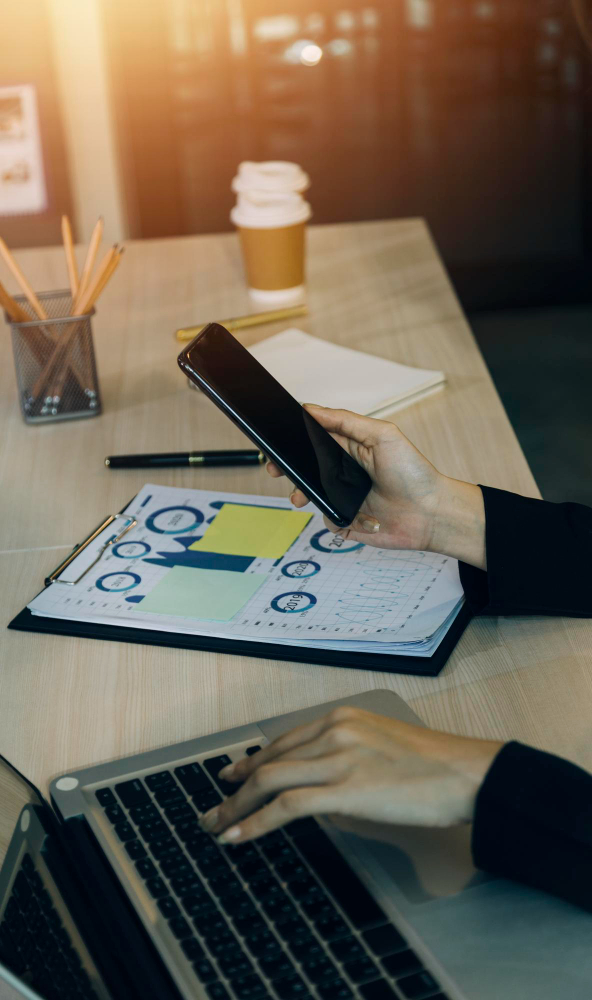 Person working on laptop while checking phone beside charts, researching a professional dating coach.