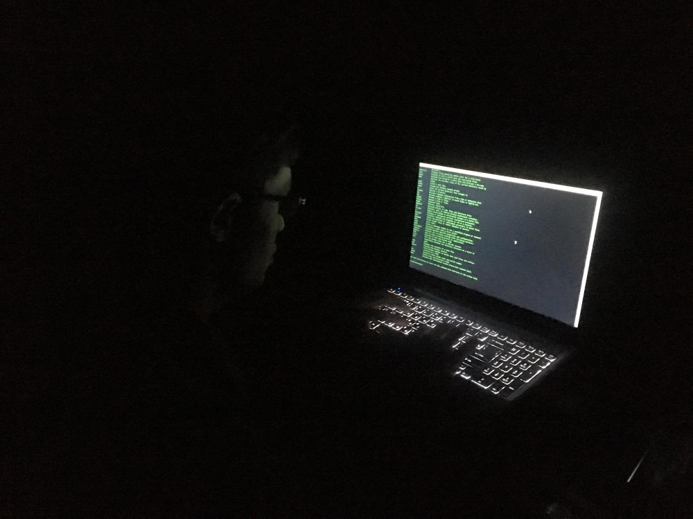 Person in dark room using a laptop with code on screen, performing dark web monitoring activities.