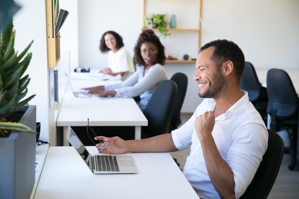 Smiling man celebrating success at laptop in office with coworkers, discussing employer risk tracking.